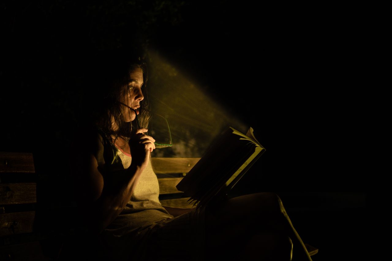 A woman reading by flashlight on a wooden bench in the nighttime.